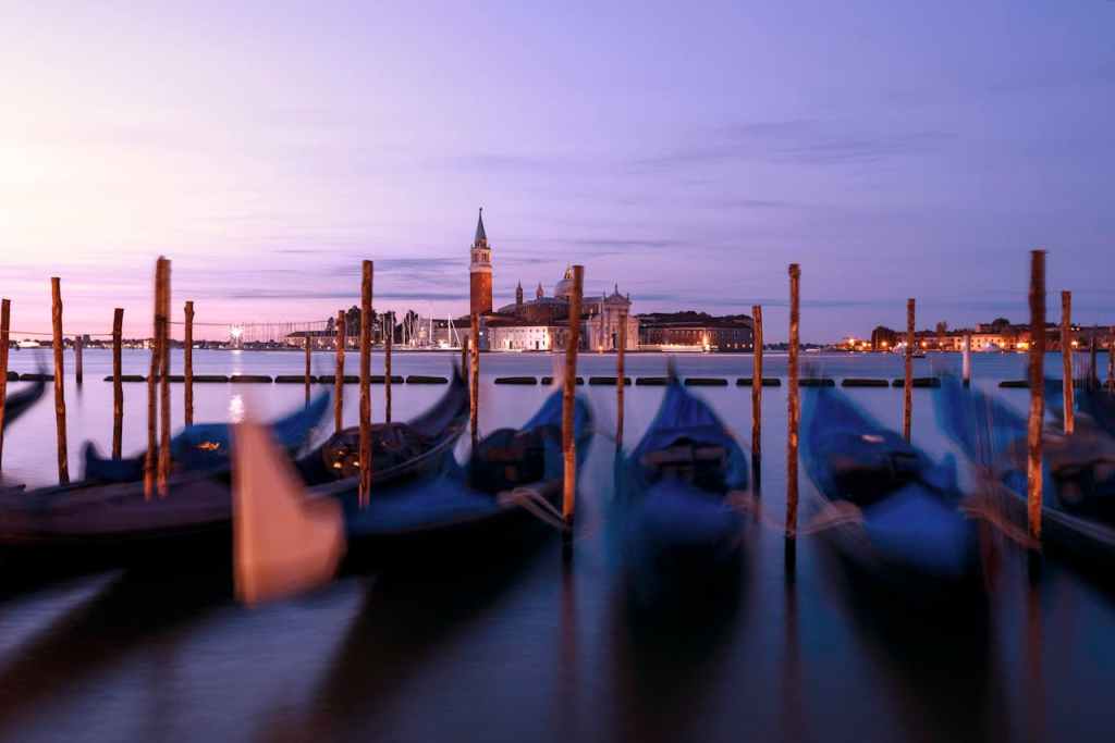 A serene view of the Venetian canals at dusk, featuring picturesque gondolas docked along wooden poles, with historic buildings and a softly lit skyline in the background.